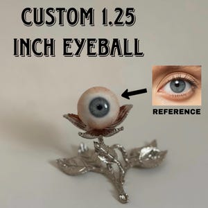 Pode incluir: Um globo ocular personalizado de 1,25 polegadas com íris azul sobre um suporte de metal prateado em forma de flor com folhas. O texto CUSTOM 1.25 INCH EYEBALL é visível no topo, incluindo uma imagem de referência de um olho humano.