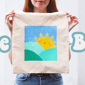 May include: A person is holding a white tote bag with a painted design of a yellow sun with a smiling face over green and blue hills. The text "Tote Bags" is in the background.
