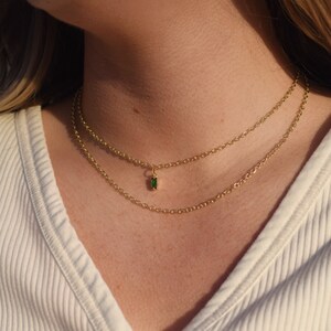 Puede incluir: Un collar de cadena de oro con un pequeño colgante de piedra preciosa verde. El collar está formado por dos cadenas.