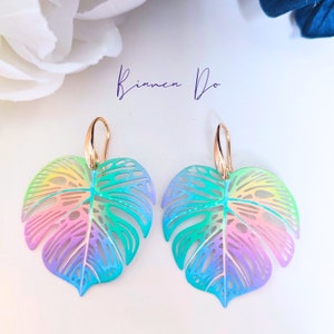 Puede incluir: Pendientes de hoja de monstera de color arcoíris con ganchos dorados. Las hojas están recortadas y tienen un diseño delicado y de encaje.