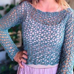 Puede incluir: Un top de crochet azul turquesa con un patrón suelto y tejido abierto. El top tiene mangas largas y un dobladillo ligeramente recortado.