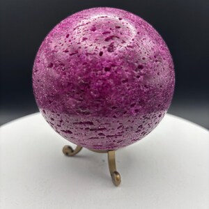 May include: A textured, deep magenta sphere, possibly a decorative stone or crystal, is displayed on a small, ornate brass stand. The sphere's surface has a pitted appearance, adding to its unique texture and visual interest. The stand has three curved legs.