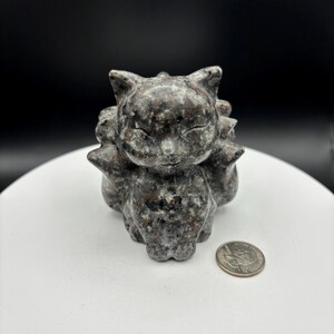 May include: A small, black and white, speckled, stone sculpture of a cat with its eyes closed. The sculpture is sitting on a white surface.