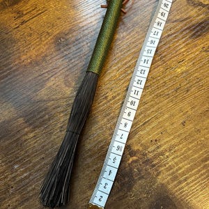 May include: A handmade whisk with a dark brown bristle head and a green wrapped handle. A leather loop is attached to the top. A white measuring tape is next to the whisk, showing inches.