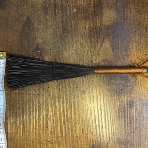 May include: A handmade brush with dark bristles and a copper-colored wrapped handle. The brush is approximately 13 inches long. A leather loop is attached to the end of the handle. The brush is lying on a wooden surface.