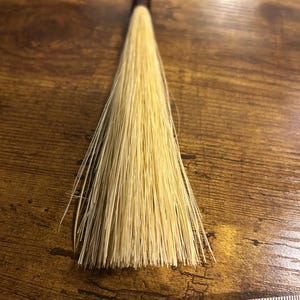 Puede incluir: Una pequeña escoba artesanal con un mango marrón oscuro y cerdas de color amarillo claro. Las cerdas están hechas de fibras naturales. Una cinta métrica con centímetros está en primer plano.