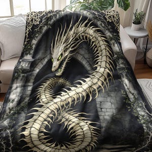 May include: A plush blanket featuring a detailed illustration of a skeletal dragon with green eyes. The dragon is set against a backdrop of a stone archway and dark, gothic-style architecture. The blanket is draped over a sofa.