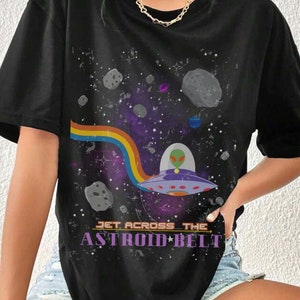 Puede incluir: Camiseta negra con un diseño espacial. Muestra un OVNI con un extraterrestre, un arcoíris y el texto "JET ACROSS THE ASTROID BELT". El fondo presenta planetas y estrellas, creando un diseño cósmico.