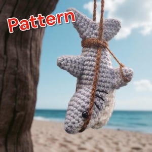 May include: A crocheted shark toy in shades of gray and white, suspended by a brown string. The shark has black stitched details and a dark eye. The word "Pattern" is displayed in red text. The background shows a beach and blue sky.