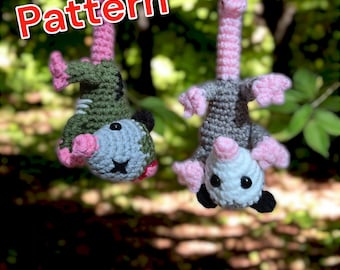 May include: Two crocheted opossum plushies hanging from a branch. One is grey and white with pink ears and nose, the other is green and grey with pink ears and nose. The word "Pattern" is written in red letters above the plushies.