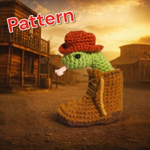 Könnte beinhalten: Eine gehäkelte Schlange mit rotem Cowboyhut, die aus einem braunen Stiefel kommt. Die Schlange ist grün mit einer rosa Zunge. Das Wort "Pattern" steht in roter Schrift oben links. Der Hintergrund ist eine Westernstadt.