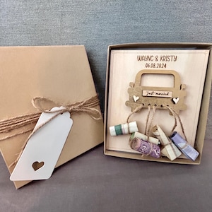 May include: A wooden box with a cutout of a car with the text "Just married" on the back window. The box is decorated with a brown ribbon and a white tag with a heart cutout. The box contains rolled up bills of money.
