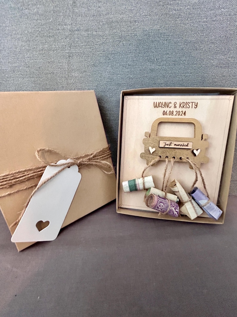 May include: A wooden box with a cutout of a car with the text "Just married" on the back window. The box is decorated with a brown ribbon and a white tag with a heart cutout. The box contains rolled up bills of money.