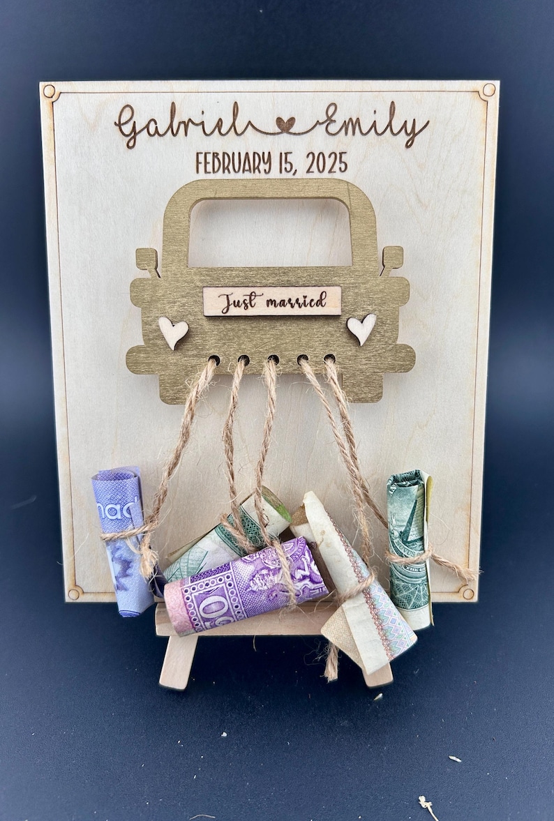 May include: A wooden sign with a cutout of a car, with the text "Just married" on the car. The sign is decorated with twine and rolled up bills. The sign is personalized with the names "Gabriel & Emily" and the date "February 15, 2025".