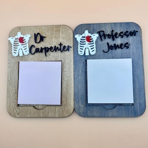 Könnte beinhalten: Zwei Holz-Notiztafeln, eine hellbraune und eine grau-blaue. Jede hat einen weißen Brustkorb mit einem roten Herzen und einen Namen: "Dr. Carpenter" und "Professor Jones". Jede Tafel hat einen quadratischen Platz für einen Haftzettel.