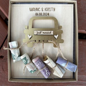 May include: A wooden box with a gold car cutout on the front. The car has a sign that says "Just married" and is decorated with hearts. The box is filled with rolled up money tied with twine.