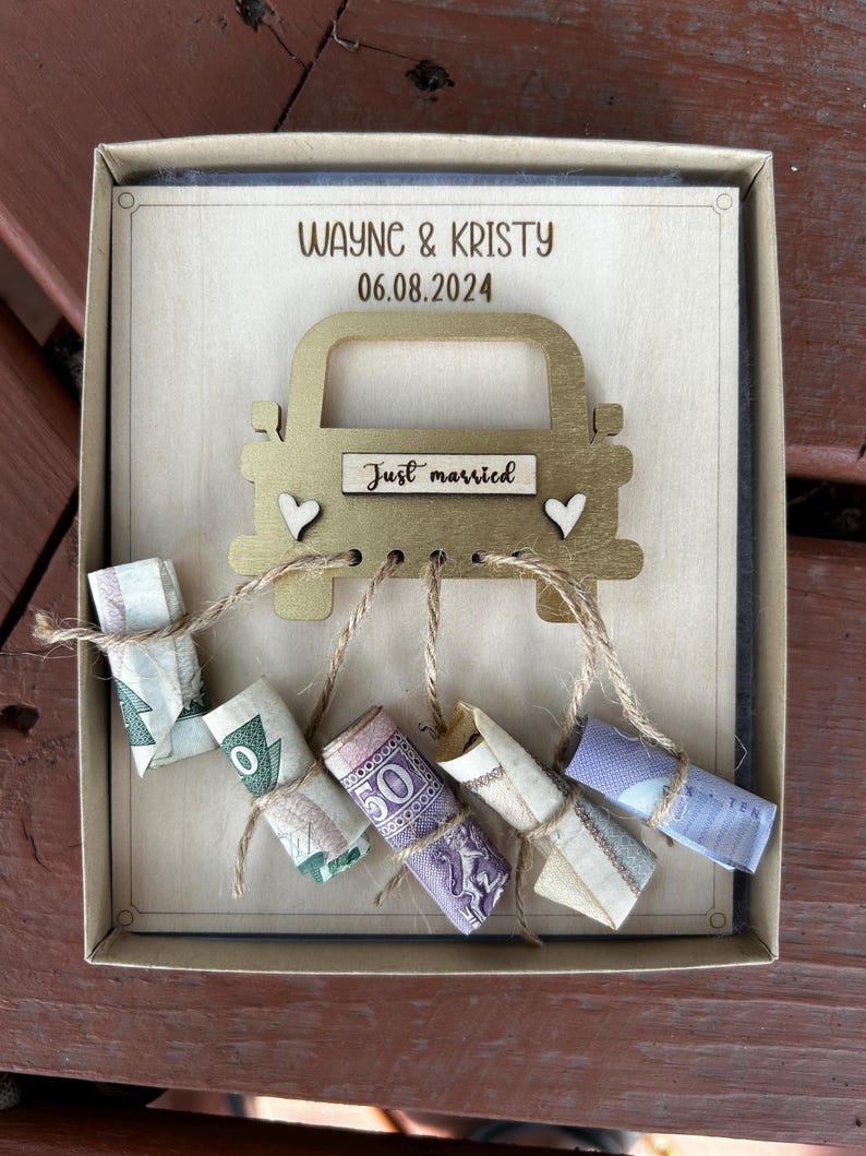 May include: A wooden box with a gold car cutout on the front. The car has a sign that says "Just married" and is decorated with hearts. The box is filled with rolled up money tied with twine.