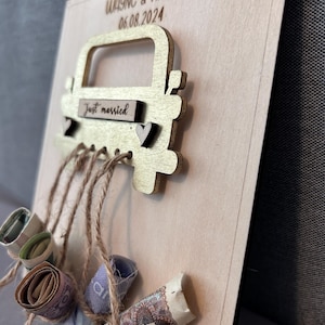 May include: A wooden card with a gold-painted car cutout attached. The car has a sign that says "Just married" and a small heart. The card is decorated with rolled-up bills tied with twine.