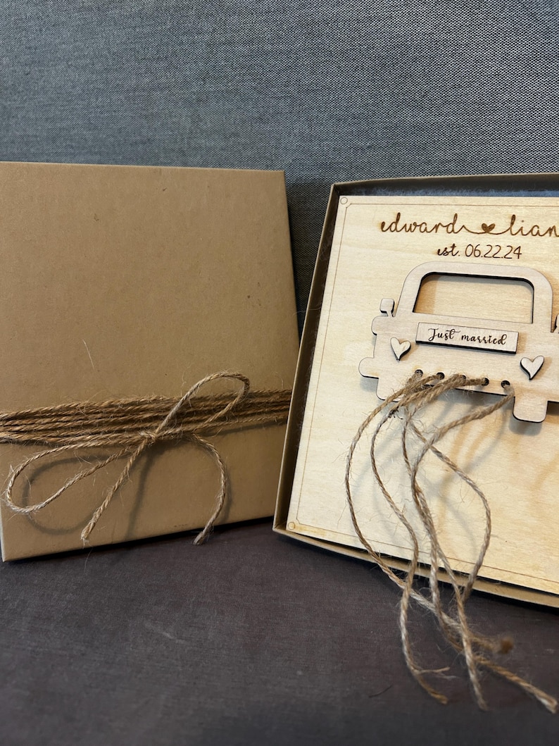 May include: A wooden box with a brown paper lid tied with twine. The box contains a wooden card with a car design and the text "Edward & Lian est. 06.22.24 Just married".