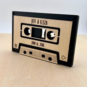 May include: A wooden cassette tape with a black border. The tape has the text "JEFF & ELIZA" and "JUNE 16, 2018" on it. The tape is a wedding keepsake.