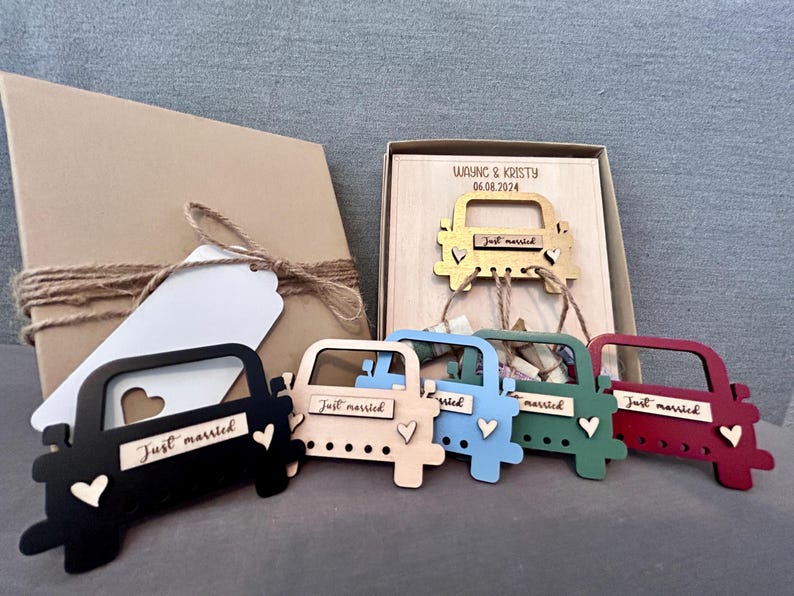 May include: Six wooden car ornaments with the words "Just Married" on them. The cars are painted in different colors: black, blue, green, red, and gold. The cars are arranged in a row on a wooden surface. The text "Wayne & Kristy 06.08.2024" is on the surface.