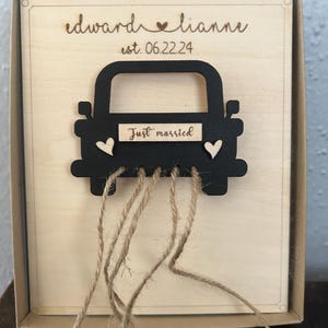 May include: A wooden box with a black silhouette of a car with the words "Just married" on the back. The car has two small hearts on the side and is attached to the box with twine. The box is engraved with the names "Edward & Lianne" and the date "est. 06.22.24".