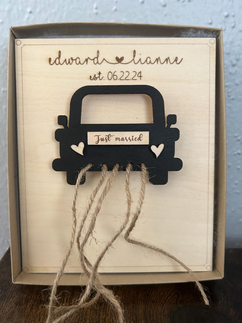 May include: A wooden box with a black silhouette of a car with the words "Just married" on the back. The car has two small hearts on the side and is attached to the box with twine. The box is engraved with the names "Edward & Lianne" and the date "est. 06.22.24".