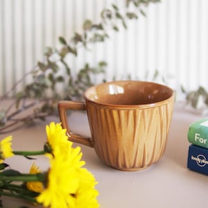 May include: A tan ceramic mug with a textured, vertical pattern. The mug has a square handle and sits next to yellow flowers and two books. The books are titled "For You When I" and "Central".
