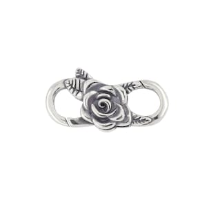 May include: A silver-colored clasp with a rose and leaf design. The clasp is oval-shaped with a spring-loaded closure. The rose is in the center of the clasp, with leaves on either side. The clasp is likely used for jewelry or other accessories.