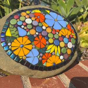 May include: A mosaic stone featuring a colorful floral design. The mosaic includes orange, yellow, and blue flowers, along with various colored circles and leaf shapes. The stone is surrounded by a dark border and set on a brick surface.