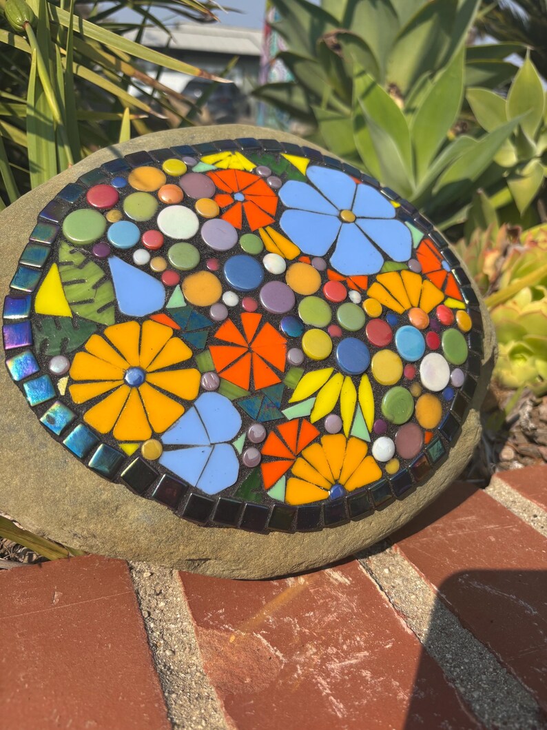 May include: A mosaic stone featuring a colorful floral design. The mosaic includes orange, yellow, and blue flowers, along with various colored circles and leaf shapes. The stone is surrounded by a dark border and set on a brick surface.