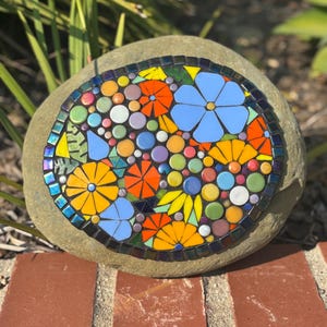May include: A mosaic stone featuring a colorful floral design. The mosaic includes blue, orange, and yellow flowers, along with various colored circles. The stone is set on a brick surface, with green foliage in the background.