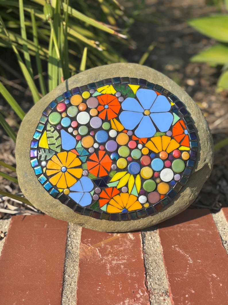 May include: A mosaic stone featuring a colorful floral design. The mosaic includes blue, orange, and yellow flowers, along with various colored circles. The stone is set on a brick surface, with green foliage in the background.