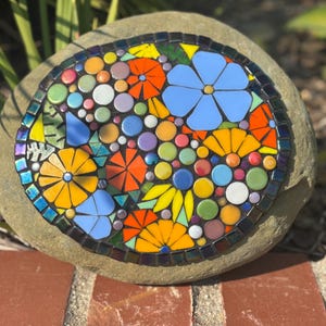 May include: A mosaic stone featuring a colorful floral design. The mosaic includes blue, orange, yellow, and white flowers, with green and purple accents. The stone is round and sits on a brick surface.