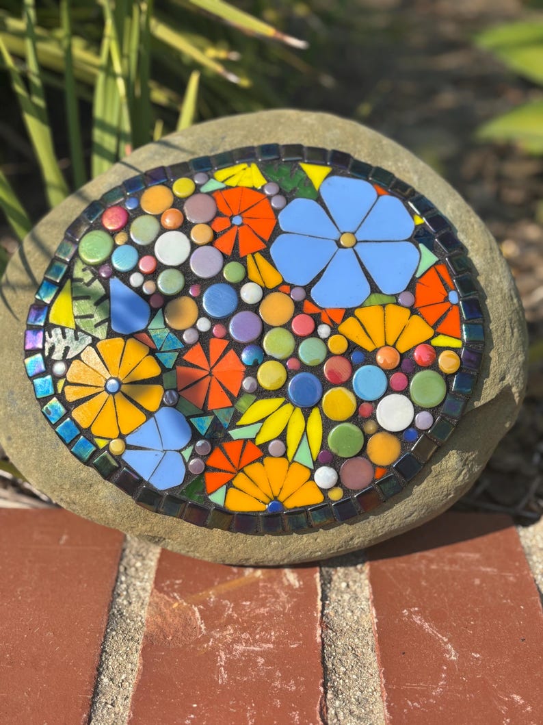 May include: A mosaic stone featuring a colorful floral design. The mosaic includes blue, orange, yellow, and white flowers, with green and purple accents. The stone is round and sits on a brick surface.