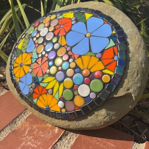 May include: A mosaic stone featuring a colorful floral design. The mosaic includes blue, orange, yellow, and green flowers, with iridescent and colorful accents. The mosaic is set on a natural, rounded stone.