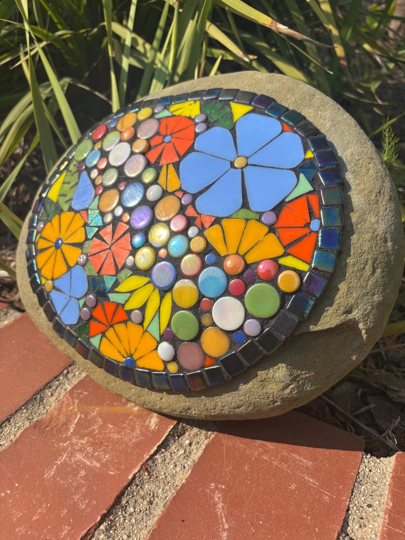 May include: A mosaic stone featuring a colorful floral design. The mosaic includes blue, orange, yellow, and green flowers, with iridescent and colorful accents. The mosaic is set on a natural, rounded stone.