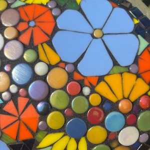 May include: A mosaic design featuring a large, light blue flower and several orange, yellow, and blue flowers. The mosaic also includes colorful, round glass pieces in various colors, including green, red, and purple. The mosaic is set against a dark background.