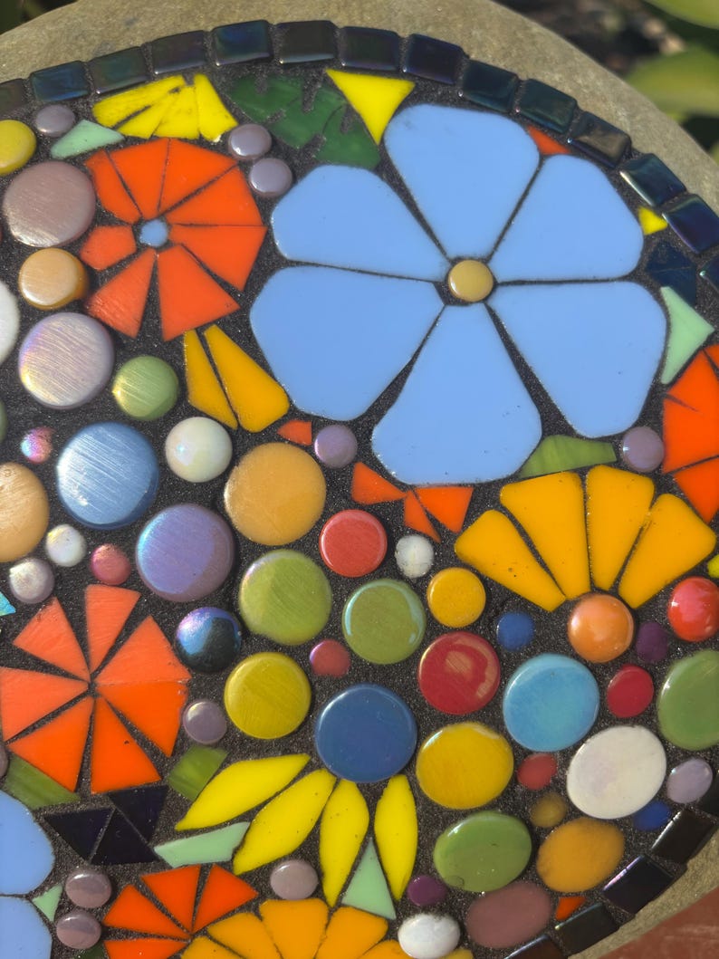 May include: A mosaic design featuring a large, light blue flower and several orange, yellow, and blue flowers. The mosaic also includes colorful, round glass pieces in various colors, including green, red, and purple. The mosaic is set against a dark background.