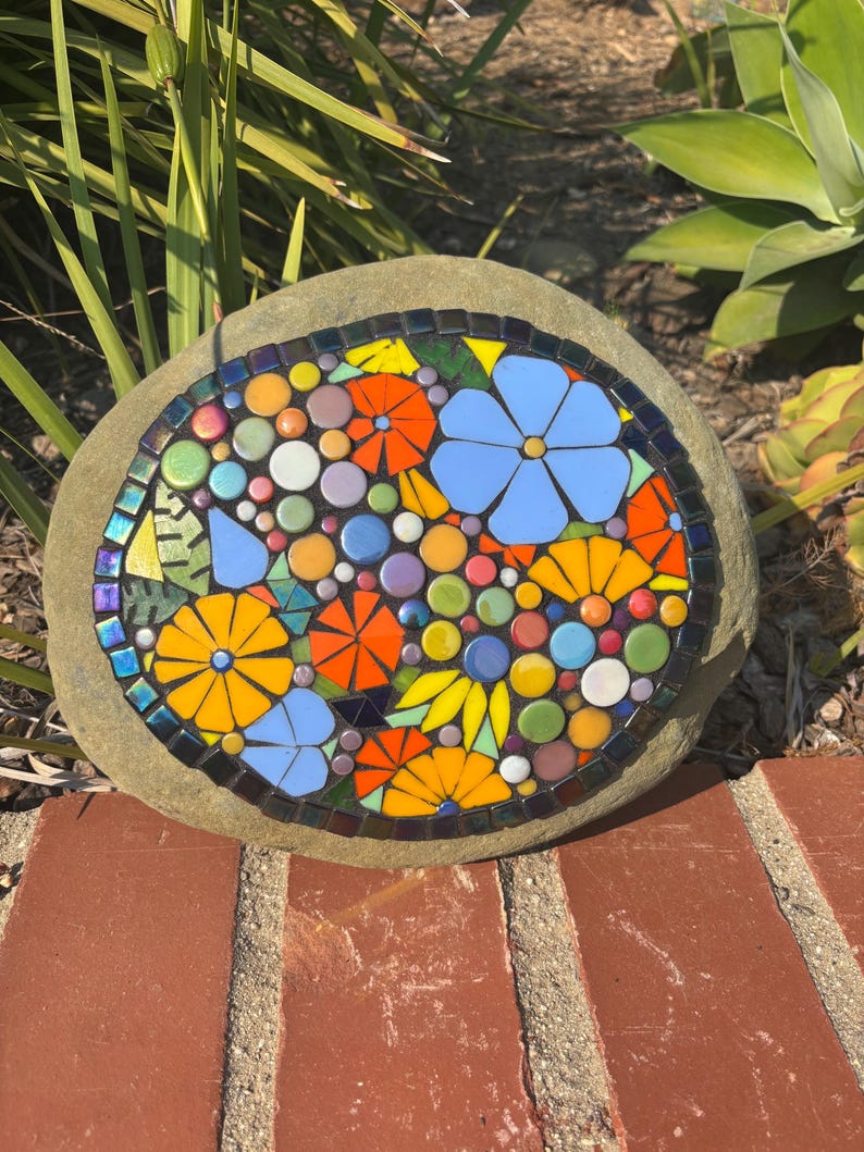 May include: A mosaic stone featuring a colorful floral design. The mosaic includes blue, orange, yellow, and white flowers, along with various colored circles. The stone is surrounded by a dark border and set against a natural background.