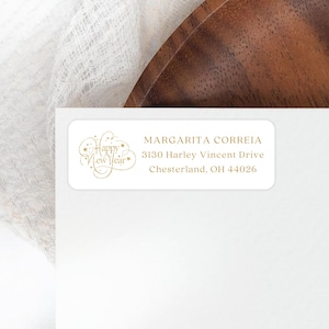 May include: A white rectangular label with gold text and a gold floral design that says "Happy New Year". The label also includes the name "Margarita Correia" and an address.