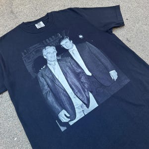 Może przedstawiać: Czarny t-shirt z szarą grafiką dw&oacute;ch mężczyzn w garniturach. Obraz jest wydrukowany na przodzie koszulki. Mężczyźni stoją blisko siebie, z jednym ramieniem na ramieniu drugiego. Koszulka ma okrągły dekolt i kr&oacute;tkie rękawy.