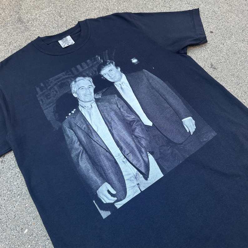 Może przedstawiać: Czarny t-shirt z szarą grafiką dw&oacute;ch mężczyzn w garniturach. Obraz jest wydrukowany na przodzie koszulki. Mężczyźni stoją blisko siebie, z jednym ramieniem na ramieniu drugiego. Koszulka ma okrągły dekolt i kr&oacute;tkie rękawy.