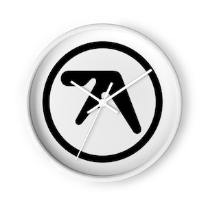 May include: A round wall clock with a white frame and a white face. The center of the clock features a black Aphex Twin logo enclosed within a black circle. The clock has three thin white hands indicating the time against the high-contrast design.