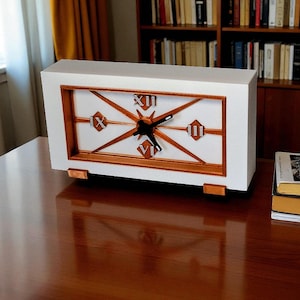 May include: A rectangular white clock with a copper-colored frame and accents. The clock face features Roman numerals and a starburst design. The clock is on a wooden desk, with books and a bookshelf in the background.