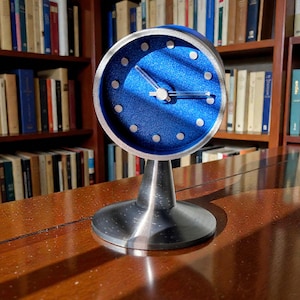 May include: A modern, silver-toned desk clock with a round, blue face. The clock features silver hour markers and hands, and sits on a sleek, conical base. The clock is displayed on a polished wooden surface, with a bookshelf in the background.