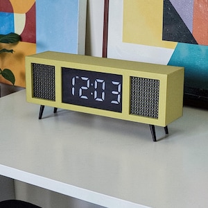 May include: A retro-style digital clock in a light yellow rectangular case with black legs. The clock face displays the time in white digits against a black background. The sides feature a speaker-like design.
