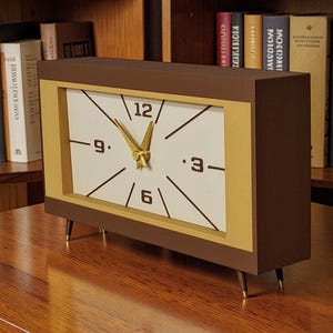 May include: A rectangular brown and gold clock with a white face. The clock has black numbers and hands, with gold-colored hands. The clock is set on a wooden surface and has four gold-tipped legs. Bookshelves are in the background.