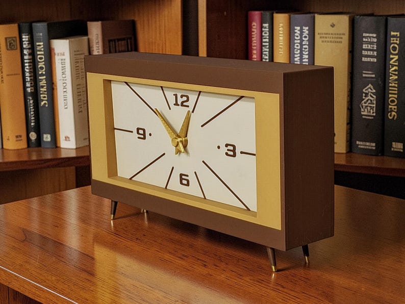 May include: A rectangular brown and gold clock with a white face. The clock has black numbers and hands, with gold-colored hands. The clock is set on a wooden surface and has four gold-tipped legs. Bookshelves are in the background.