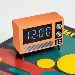 May include: Orange retro-style digital clock shaped like a vintage television set. The clock displays the time 12:00 in white digits on a black screen. The clock has black legs and decorative silver-colored knobs.